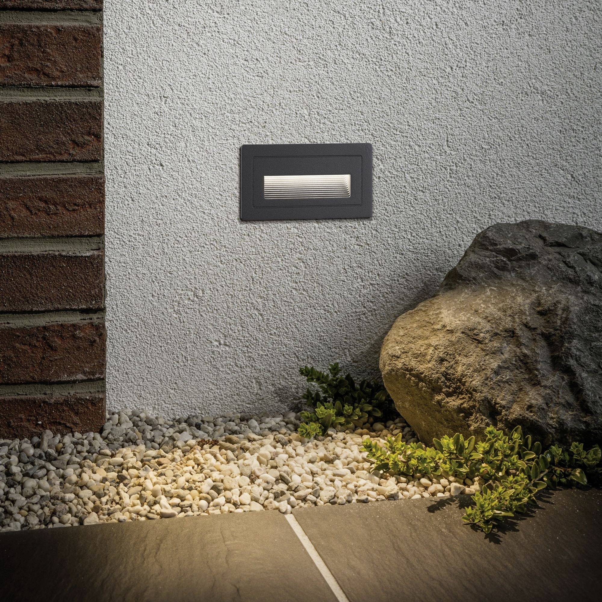 Un luminaire LED extérieur encastré dans un mur éclaire discrètement le sol composé de galets et de dalles, à côté d'une plante de pierre.