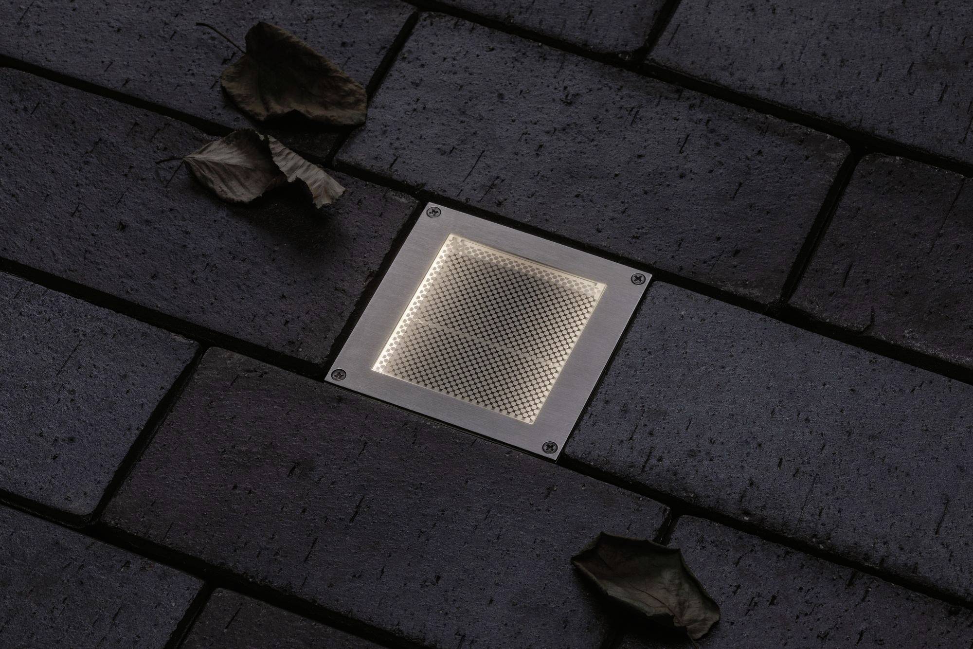 Projecteur de sol illuminé dans un pavage sombre, entouré de quelques feuilles sèches.