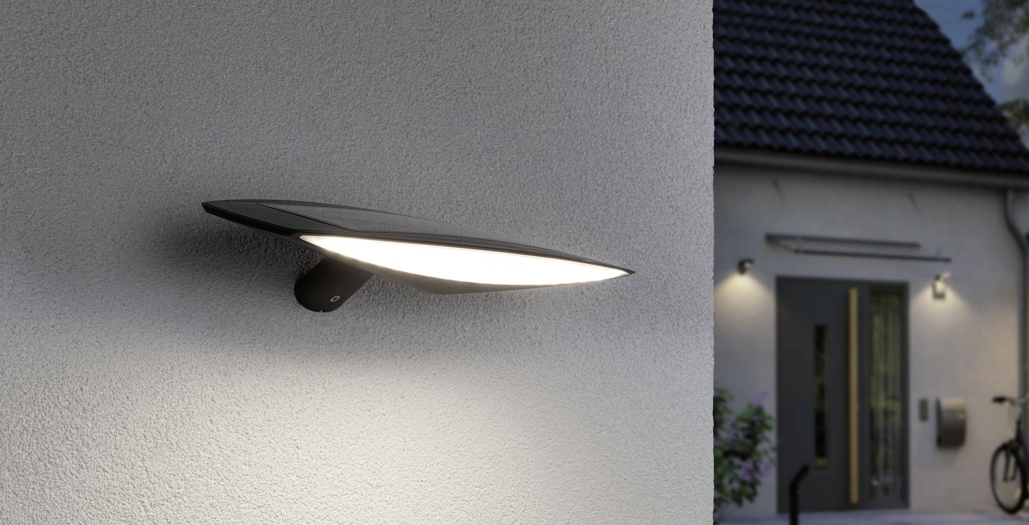 Une lampe extérieure moderne sur un mur de maison éclaire l'entrée d'une maison au crépuscule. Une porte est visible en arrière-plan.
