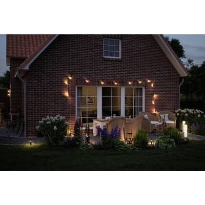 Maison de briques rouges au crépuscule, avec une terrasse éclairée, des meubles de jardin et des plantes en fleurs au premier plan.
