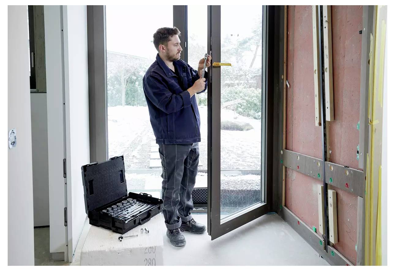 Un homme installe une grande porte en verre, en utilisant une trousse d'outils placée à proximité. La pièce est en construction, avec des murs exposés et des fenêtres.