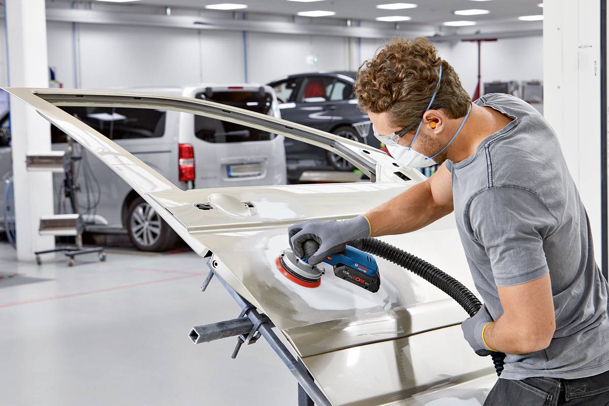 Une personne utilise un outil de meulage pour travailler la surface métallique brillante d'une portière de voiture dans un atelier. Des voitures sont visibles en arrière-plan.