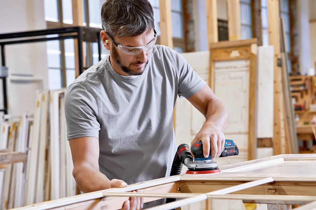 Un homme utilise une ponceuse électrique pour lisser du bois dans un atelier. Il porte des lunettes de protection et se concentre sur son travail.