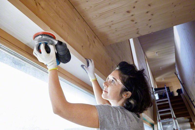 Une femme ponce un bord en bois dans une maison moderne. Elle porte des gants de protection et des lunettes de sécurité. Scène d'intérieur avec une échelle en arrière-plan.