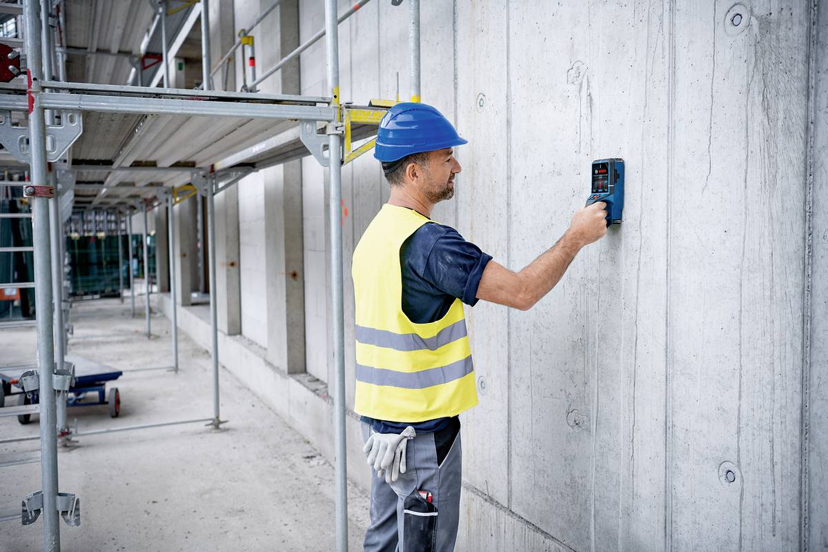 Un ouvrier portant un gilet jaune et un casque bleu utilise un appareil de mesure sur un mur en béton sur un chantier de construction équipé d'échafaudages.