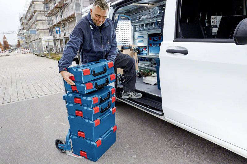Un homme décharge une pile de boîtes à outils bleues avec des fermetures rouges d'un fourgon. Un chantier de construction est visible en arrière-plan.