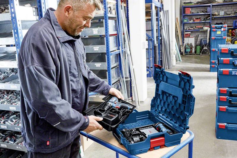 Un homme ouvre une valise à outils dans un entrepôt. La valise contient des outils électriques. Des étagères en arrière-plan sont remplies d'autres outils.