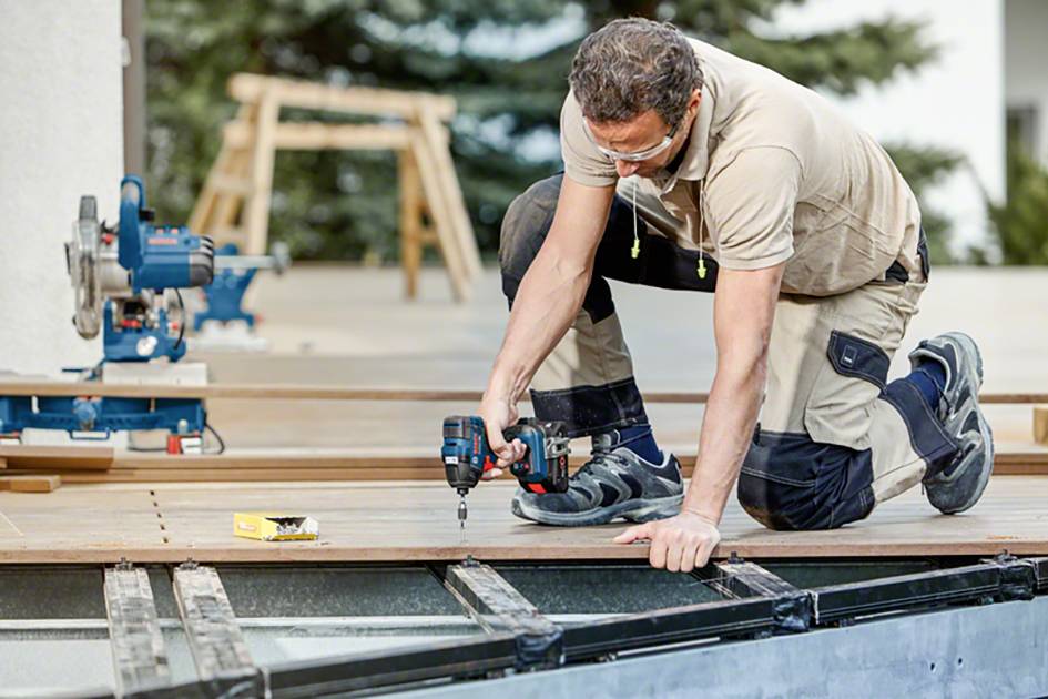 Un homme travaille sur un chantier, est à genoux sur un pont en bois et utilise une visseuse sans fil pour fixer une planche de bois.
