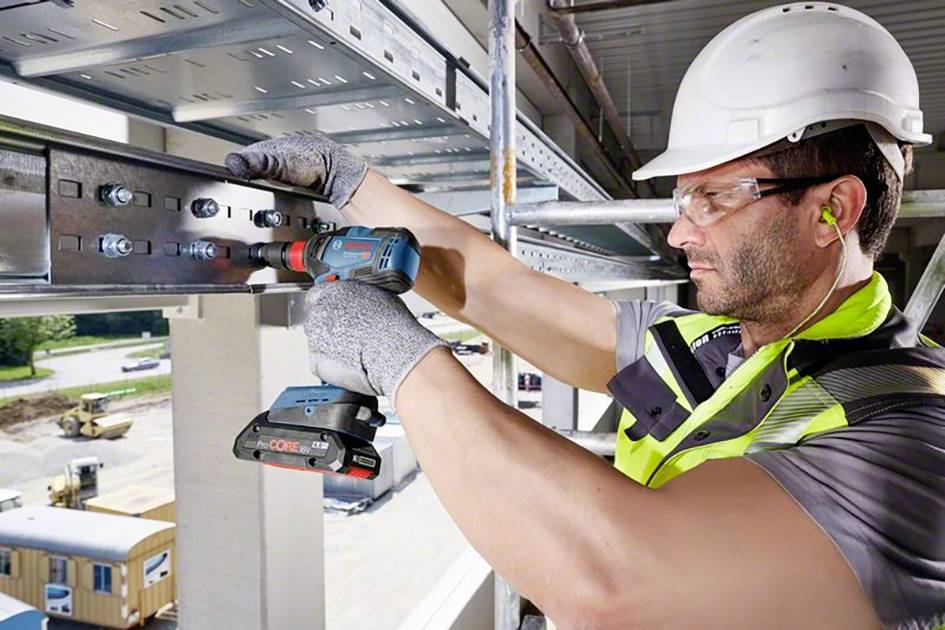 Un ouvrier portant un casque de chantier et des gants installe des poutres métalliques à l'aide d'une perceuse électrique sur un chantier de construction.