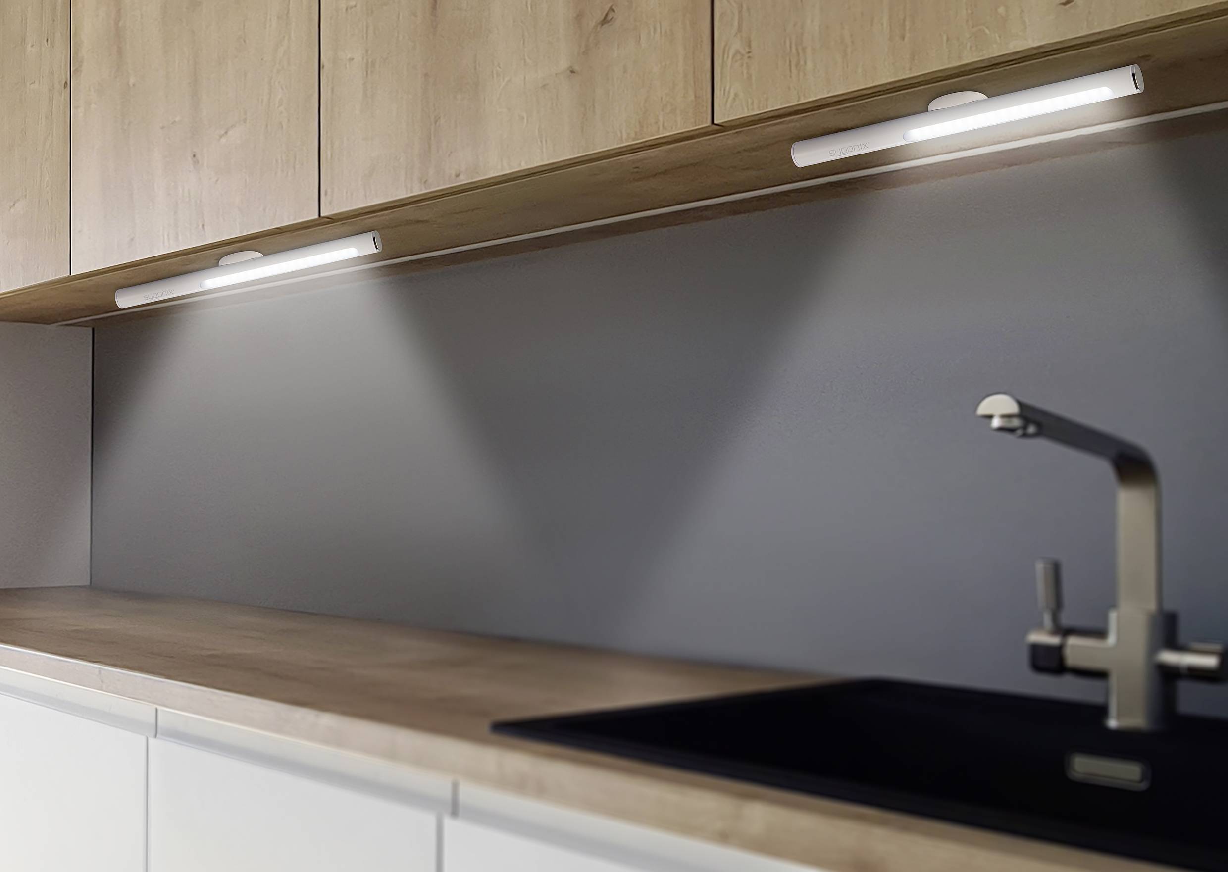 Cuisine intérieure avec des armoires en bois modernes, éclairée par deux luminaires sous-meuble, crédence grise et robinetterie en acier inoxydable au-dessus d'un évier noir.