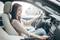 Une femme est assise au volant d'une voiture, souriante, et manipule le tableau de bord. Elle porte une chemise claire et un jean.
