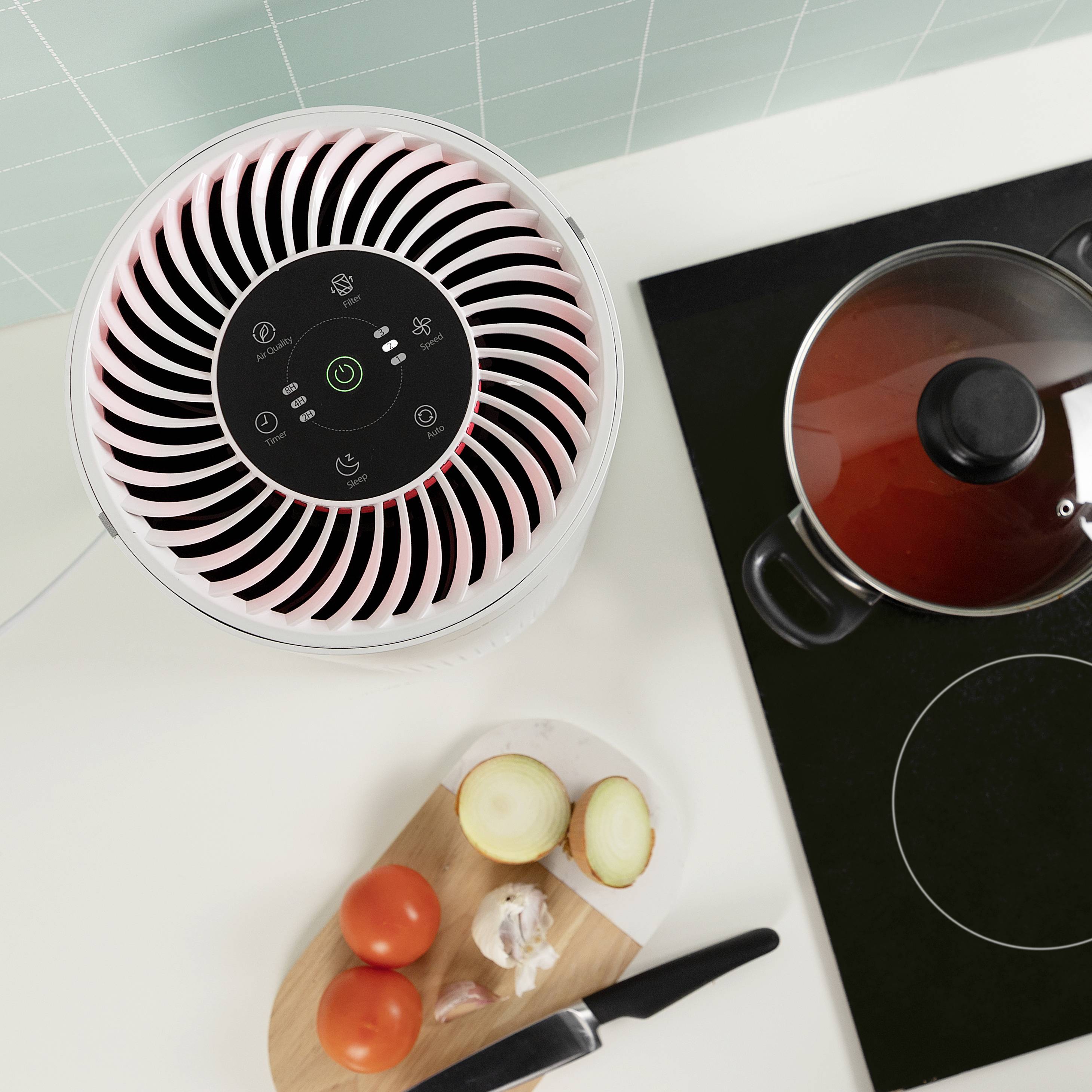Un purificateur d'air est placé à côté d'une cuisinière avec une casserole et une planche à découper contenant des tomates, des oignons et de l'ail sur un plan de travail de cuisine.