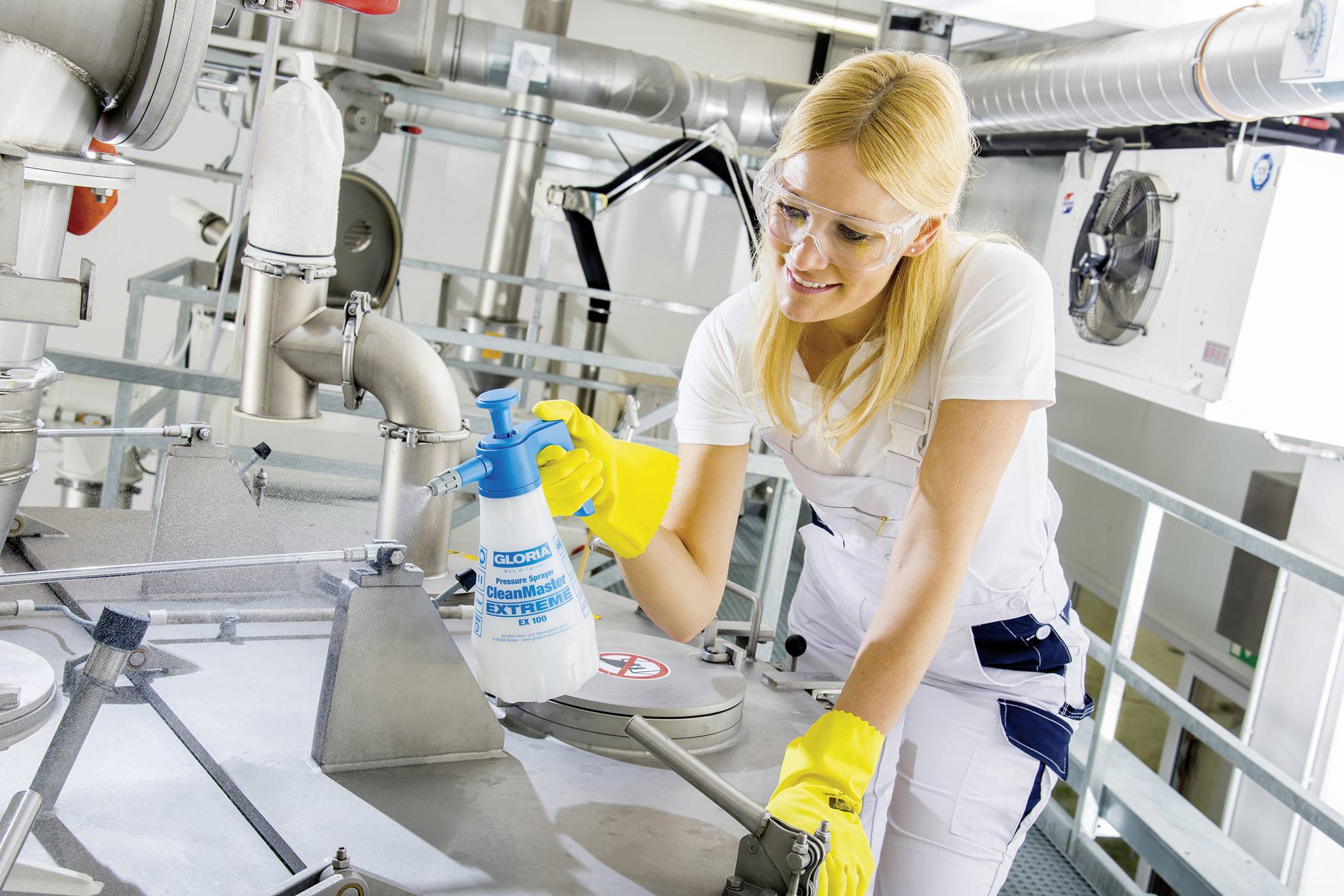 Une femme en tenue de protection et gants nettoie des machines industrielles avec un produit pulvérisé dans un environnement d'usine.