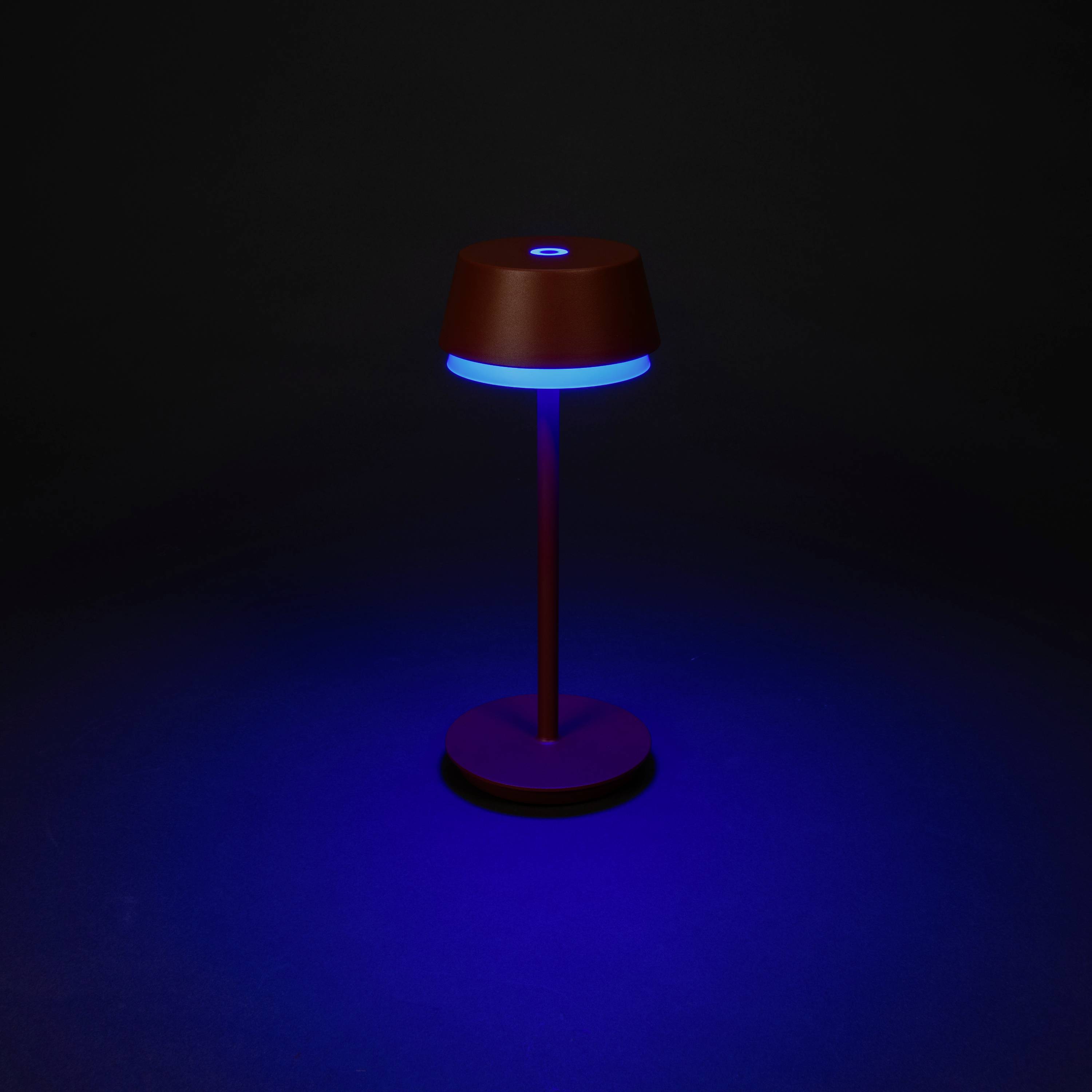 Lampe avec support rouge et anneau lumineux bleu, sur un fond sombre. Elle projette une lumière bleue sur le sol.