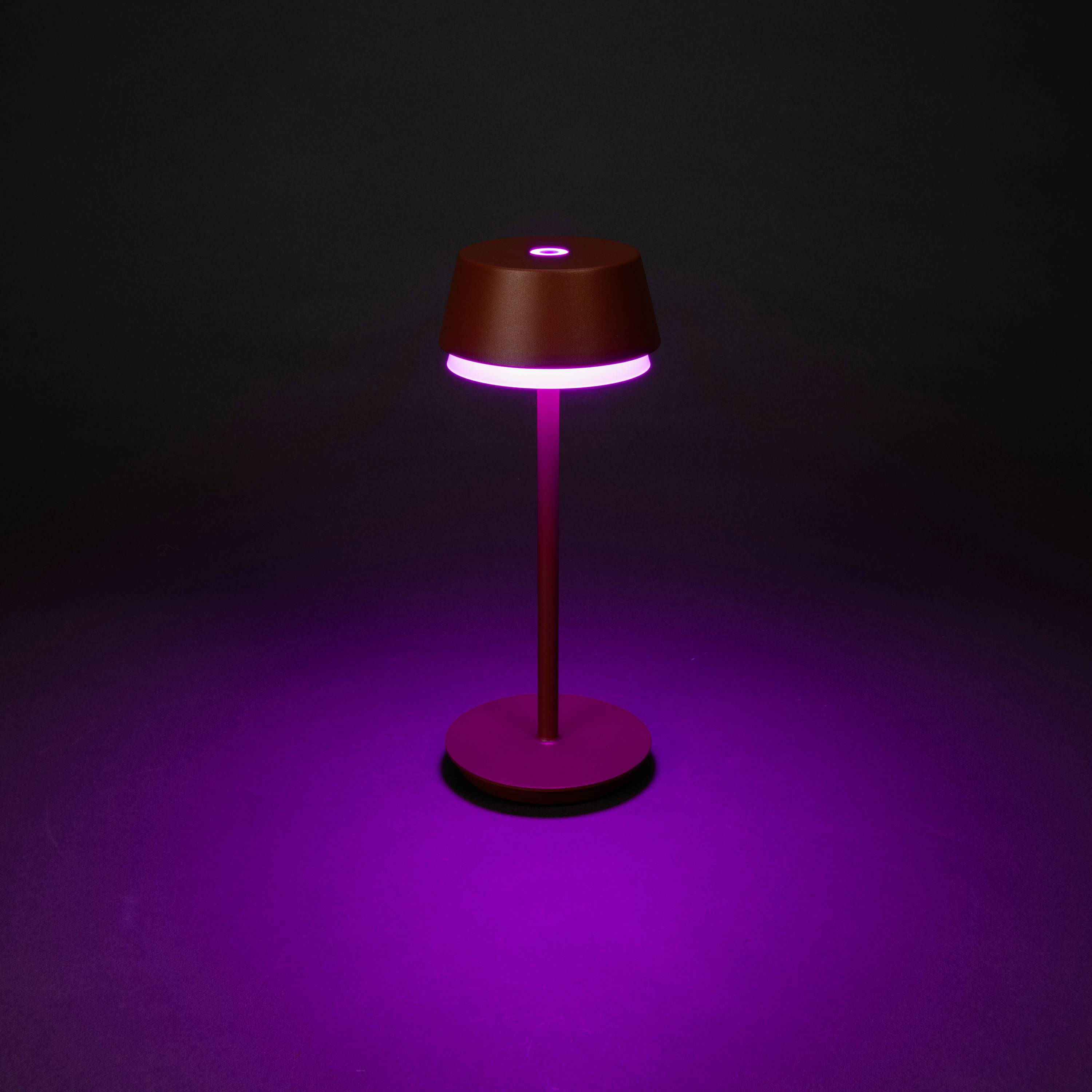Une lampe de table rouge et rose diffuse une lumière violette et se trouve sur un fond sombre.