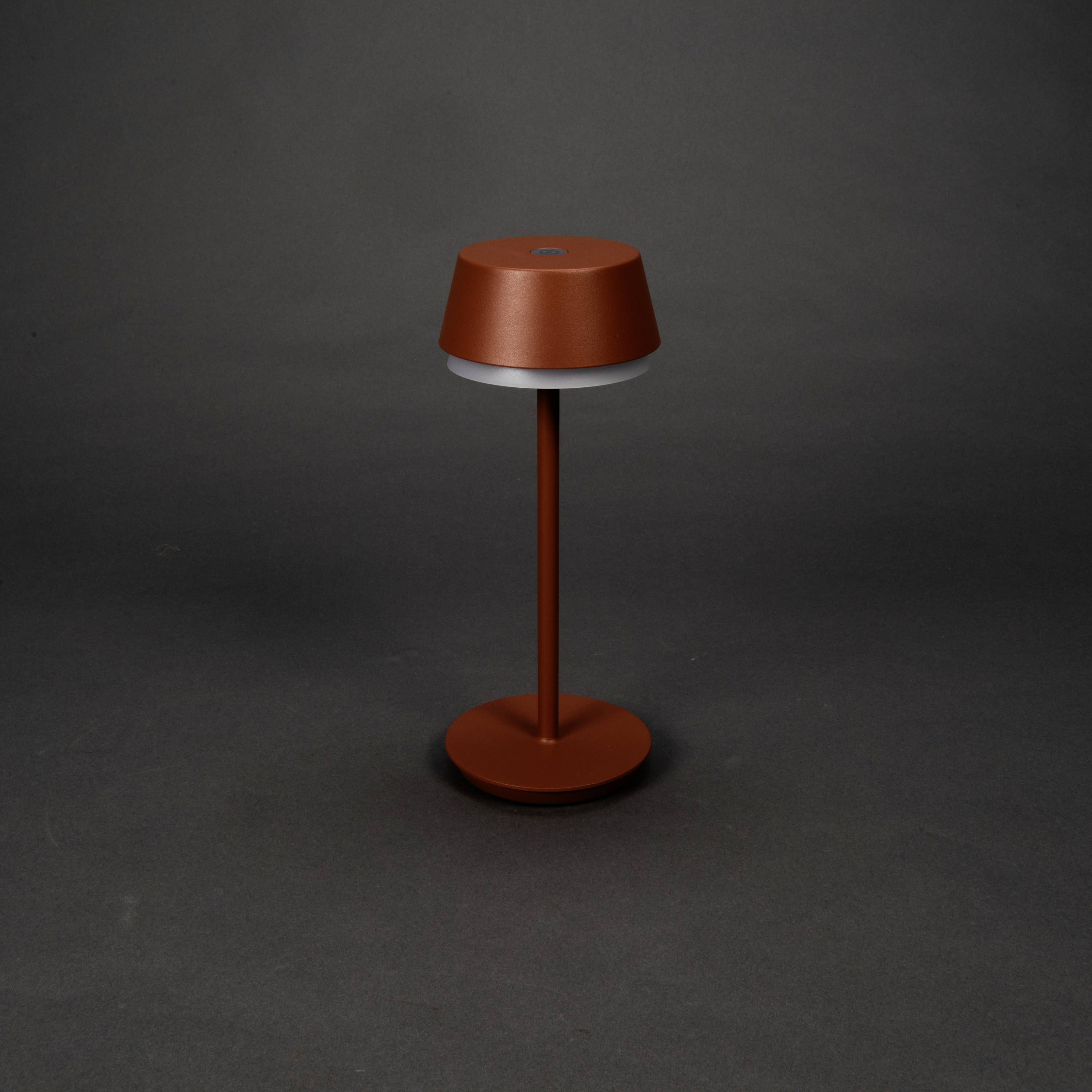 Une lampe de table moderne, rouge, avec un abat-jour cylindrique et un support fin, est posée sur un fond gris.