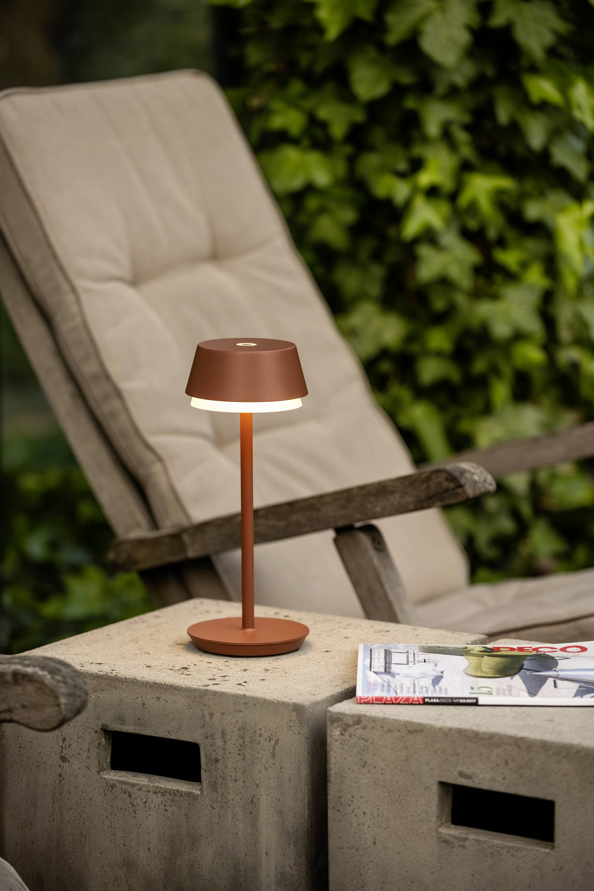 Une lampe de table marron s'allume sur un bloc de béton à côté d'un transat à l'extérieur. Un magazine est posé à proximité. En arrière-plan, on aperçoit des feuilles vertes.