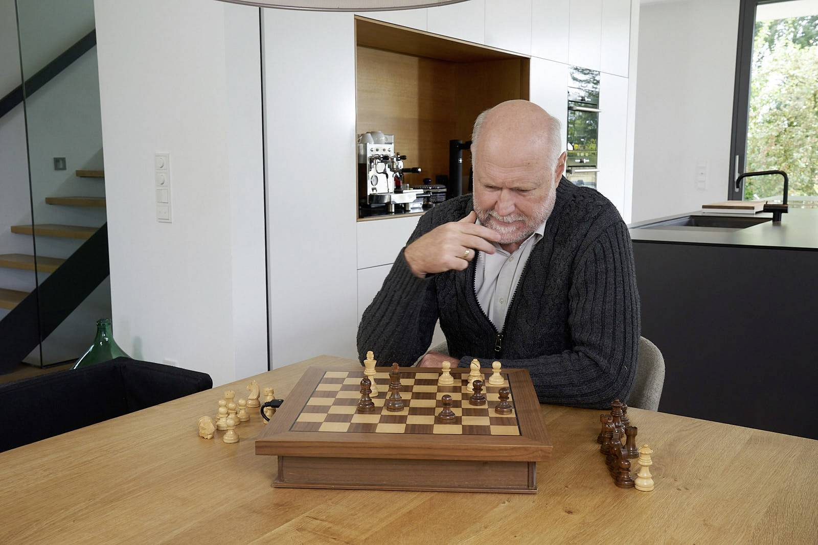Un homme âgé est assis pensivement à une table et joue aux échecs dans un salon moderne. Derrière lui, on aperçoit une cuisine et un escalier.