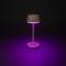 Une lampe de table moderne illumine un espace avec une lumière violette, entourée de pénombre. La lampe possède un socle rond et un abat-jour conique.