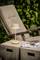 Une lampe de table beige est posée sur un bloc de pierre à côté d'un magazine. En arrière-plan, un fauteuil rembourré en bois et des plantes vertes.