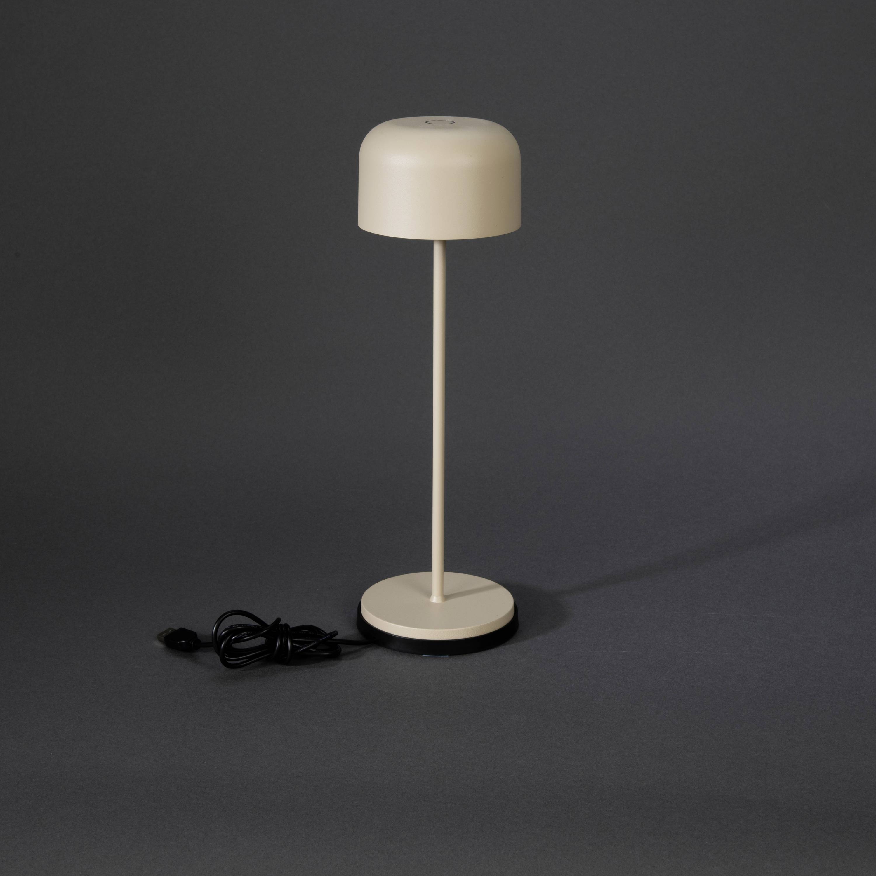 Une lampe de table beige au design simple et moderne, avec un pied long et fin. Un cordon noir décontracté est posé à côté.