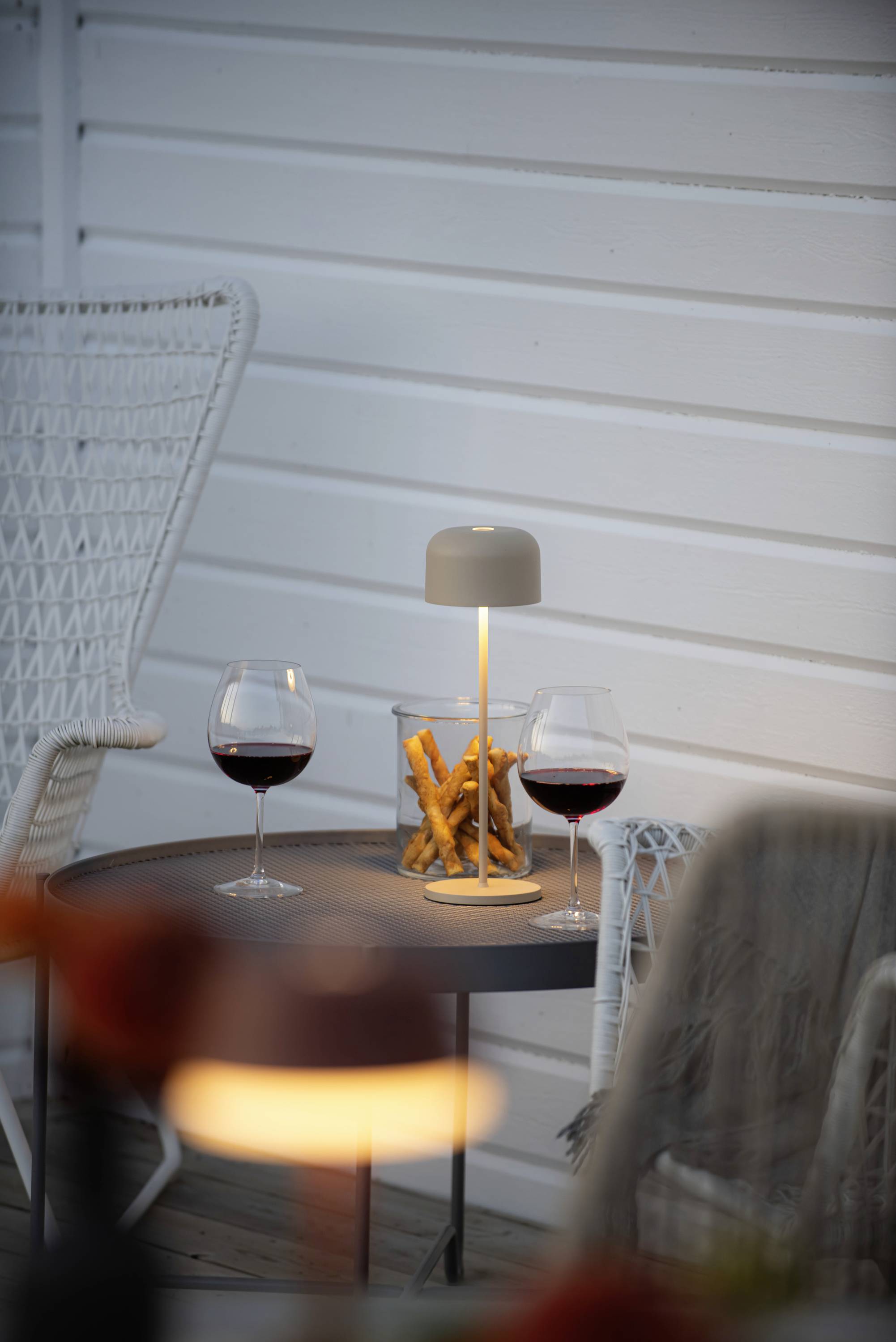 Une table avec deux verres de vin rouge, une petite lampe et un verre de gressins, sur une terrasse avec un mur en bois blanc.