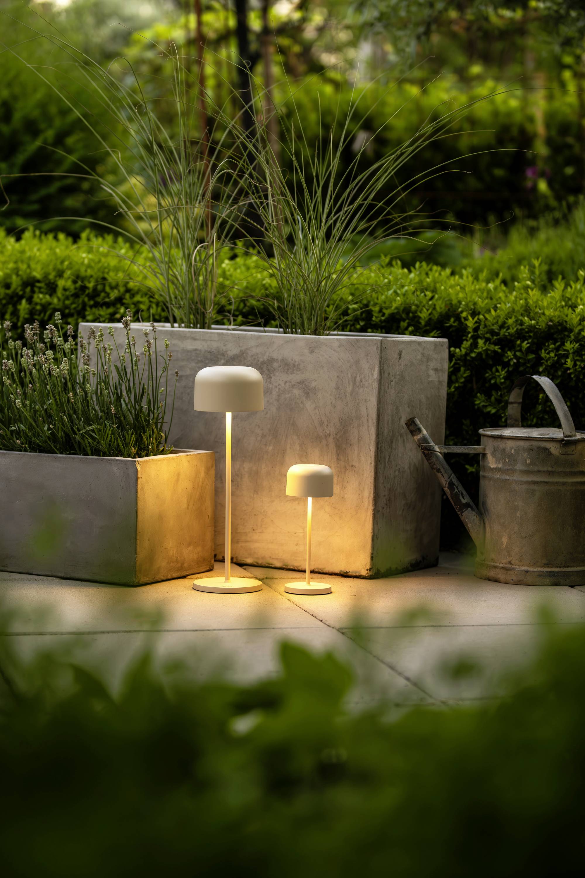 Deux lampadaires modernes et lumineux dans un jardin, entourés de plantes vertes et d'un arrosoir. Ils diffusent une lumière chaleureuse.