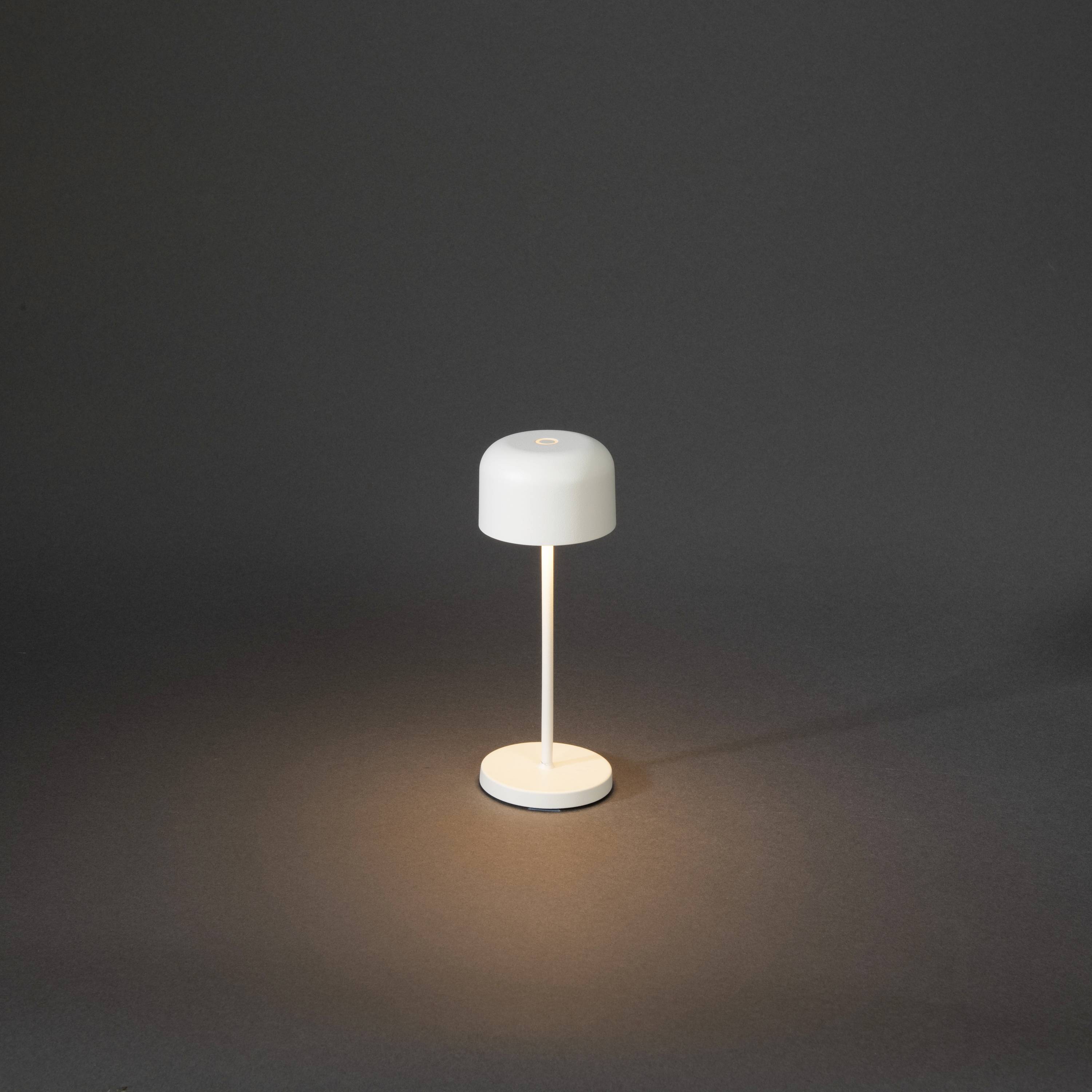 Lampe de table blanche avec abat-jour rond et pied fin, émettant une faible lueur dans un environnement sombre. Design minimaliste.