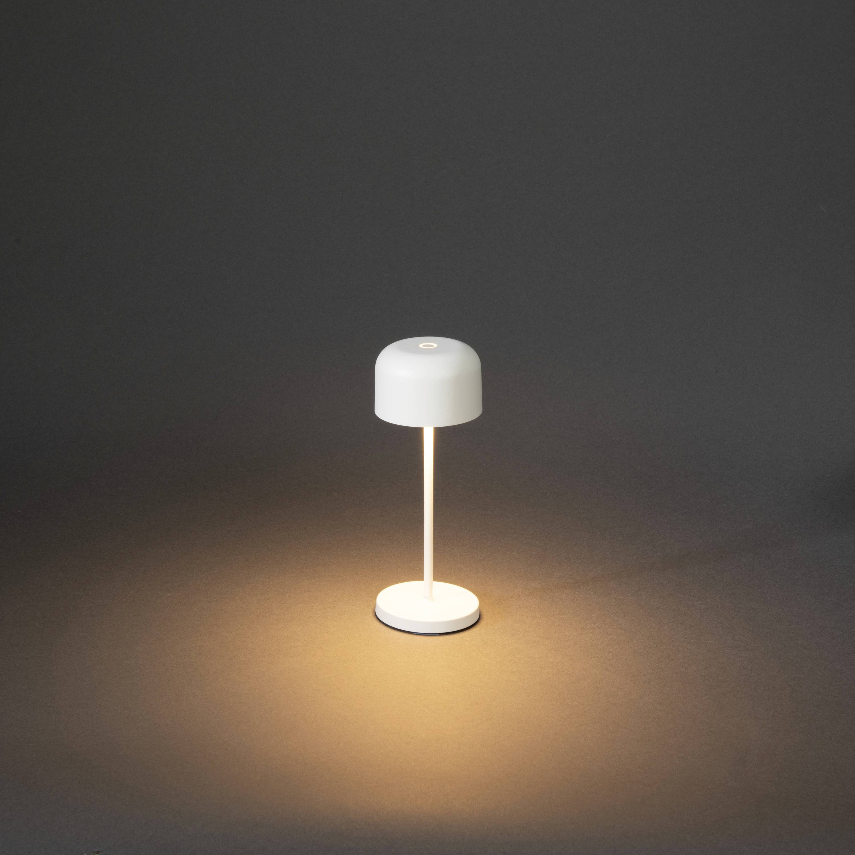 Une lampe de table moderne à abat-jour et base blancs diffuse une lumière chaleureuse. Elle se trouve dans une pièce sombre et éclaire le sol.