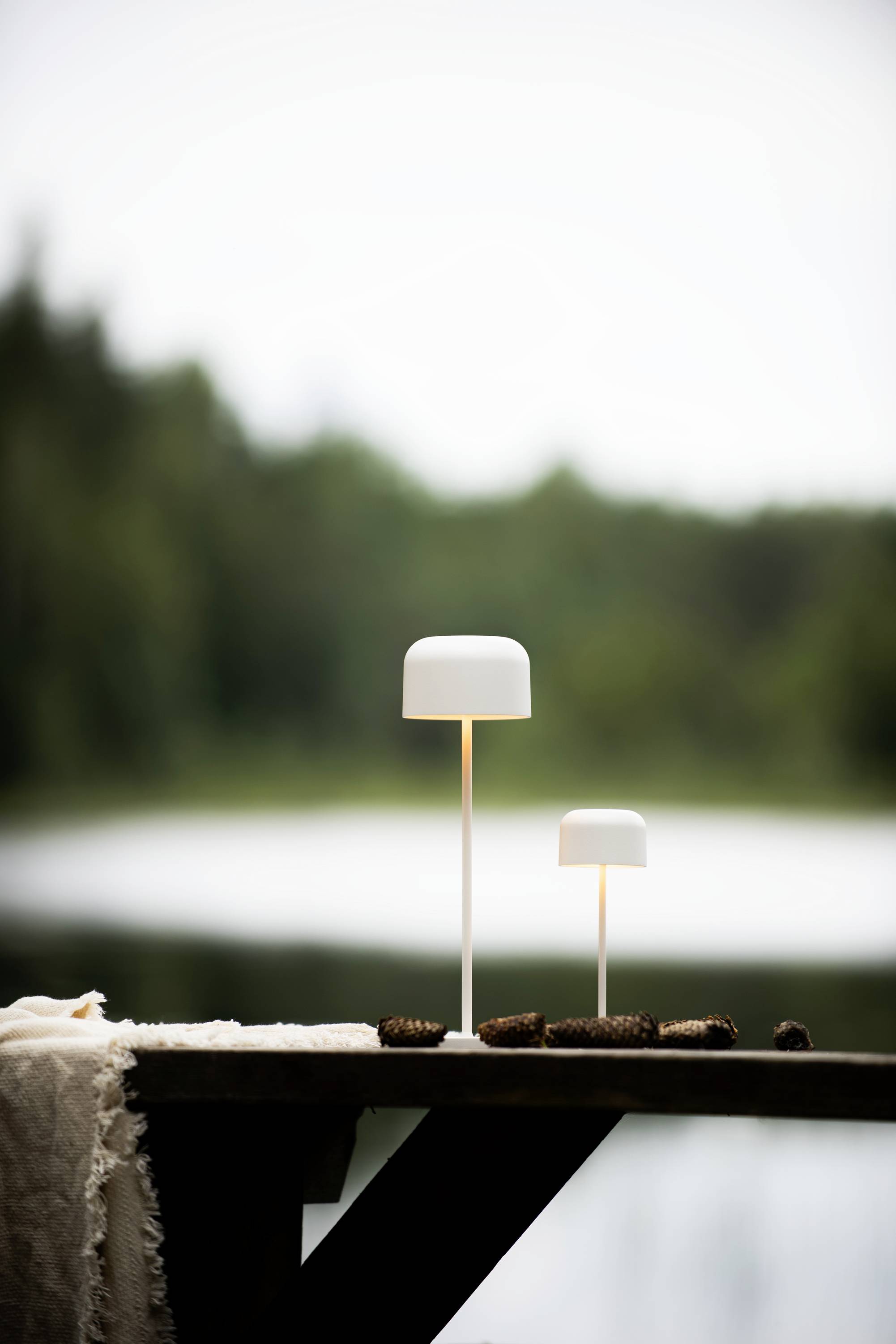 Deux lampes de table blanches de tailles différentes sur une table en bois, avec une vue sur un lac et une forêt flous en arrière-plan.