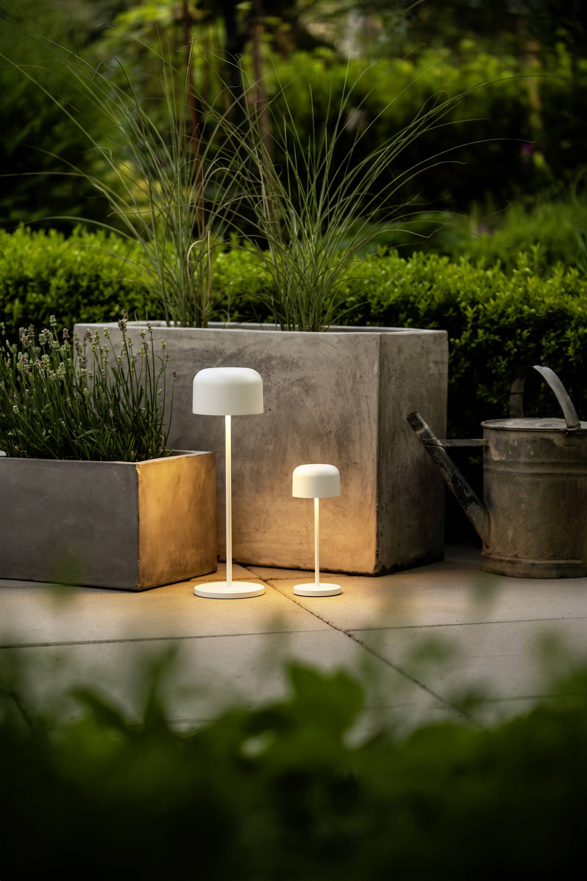 Deux lampes de table modernes brillent à l'extérieur, à côté de jardinières et d'un arrosoir, entourées de feuillage vert.