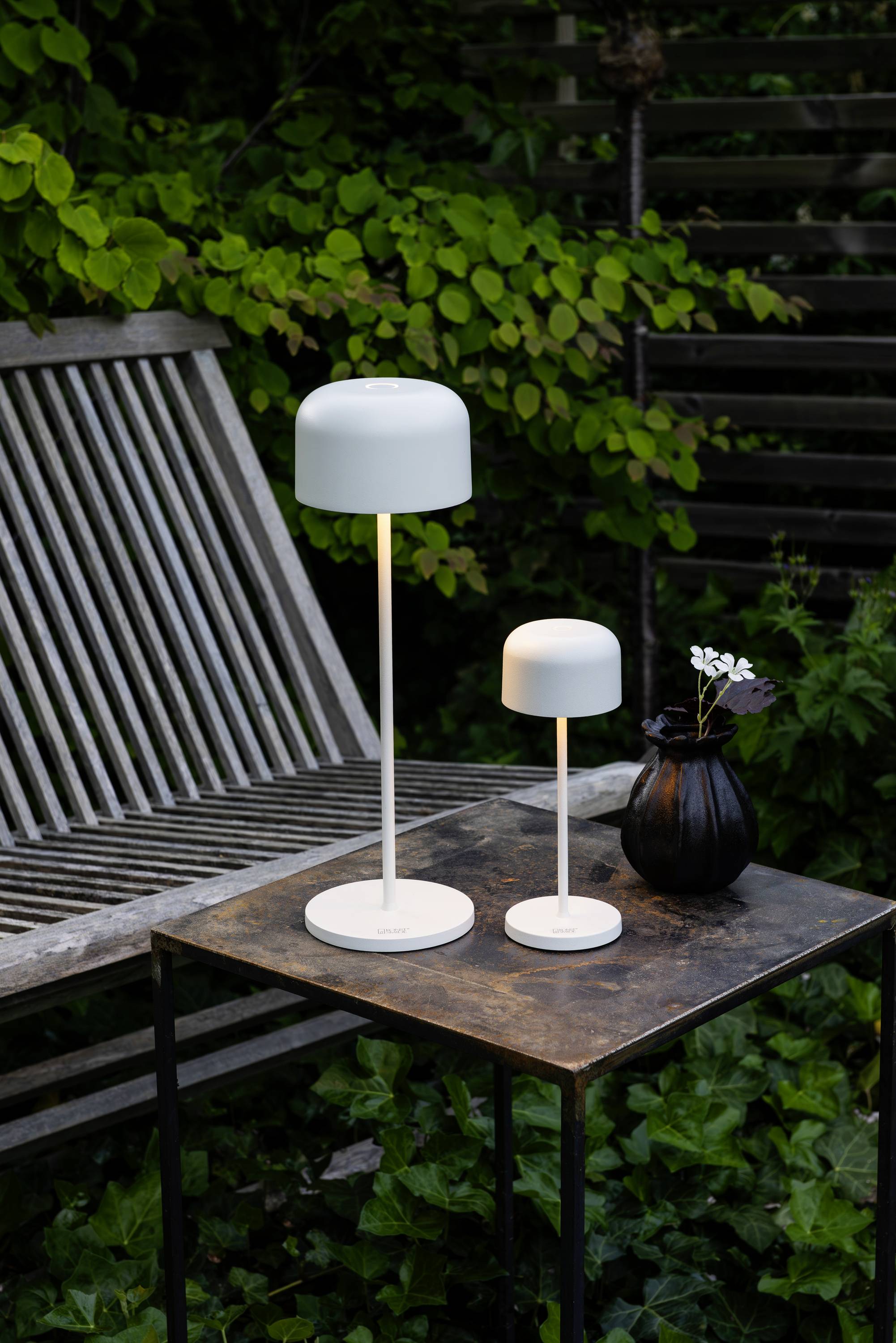 Deux lampes de table blanches de tailles différentes sont posées sur une table en métal. À côté, un vase noir contenant des fleurs, le tout devant un feuillage vert et un banc en bois.