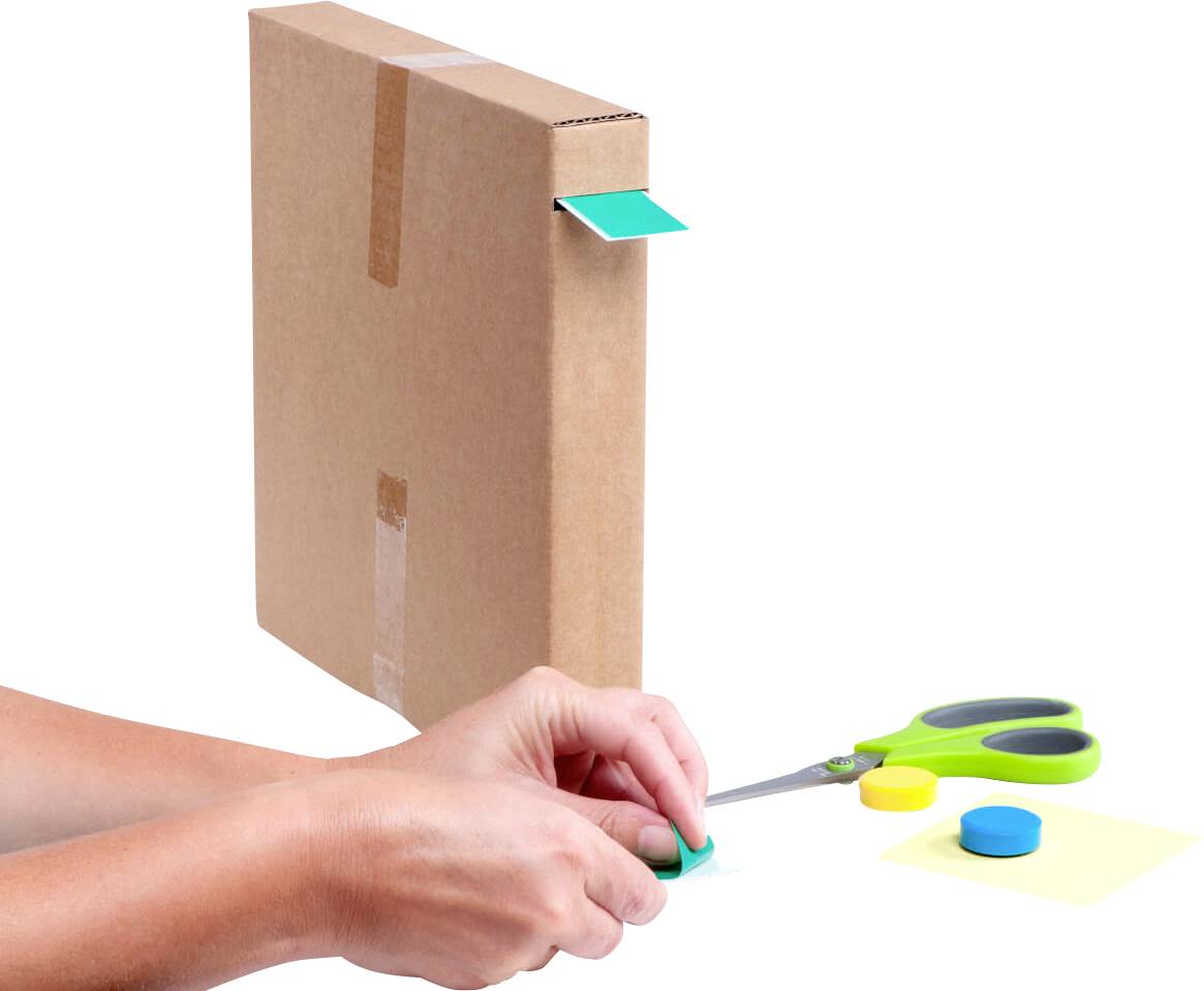 Des mains collent un aimant bleu sur une bande de papier verte qui dépasse d'un carton debout et emballé ; à côté, une paire de ciseaux verte.