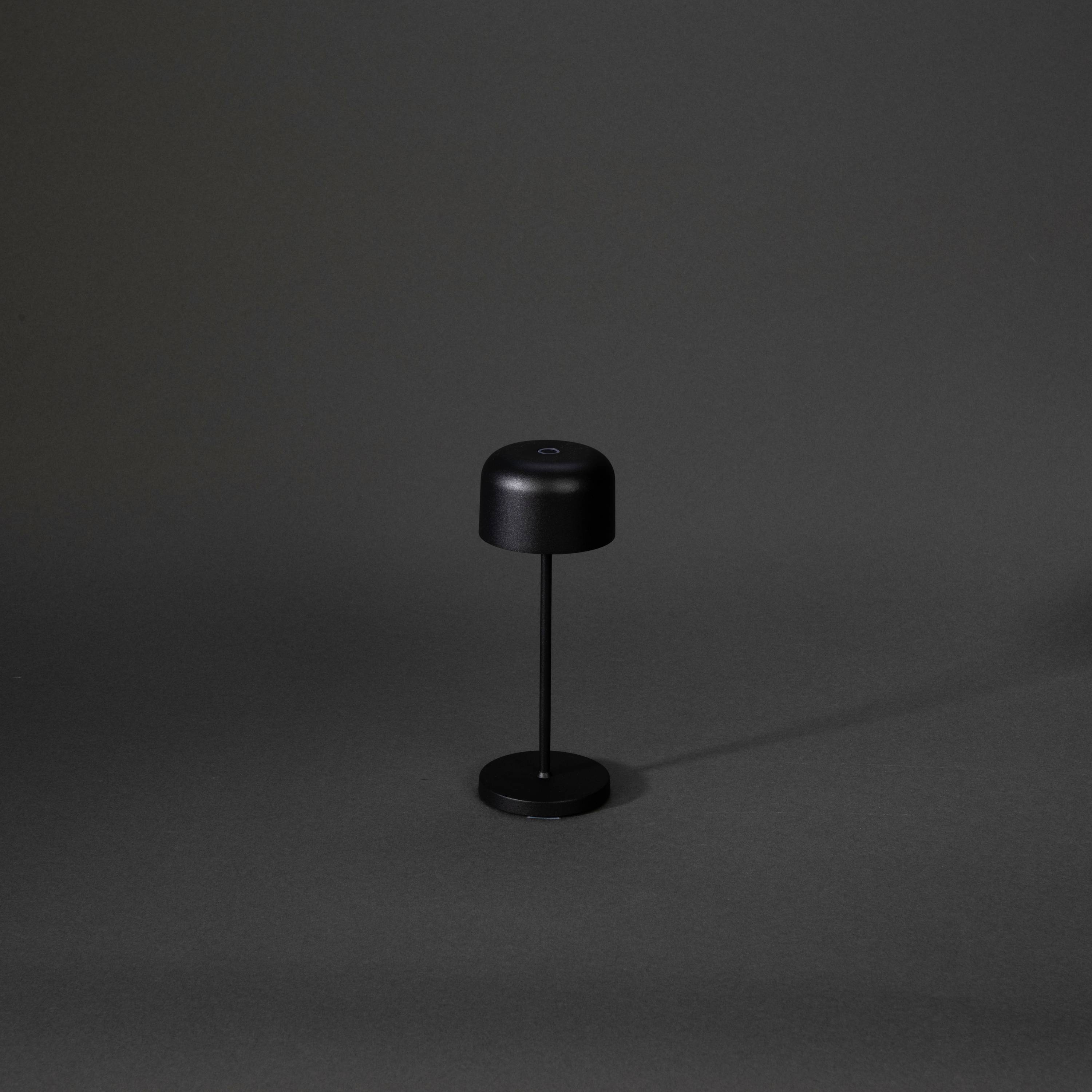 Petite lampe de table noire avec un abat-jour rond sur un pied étroit, placée devant un arrière-plan sombre. Design minimaliste.