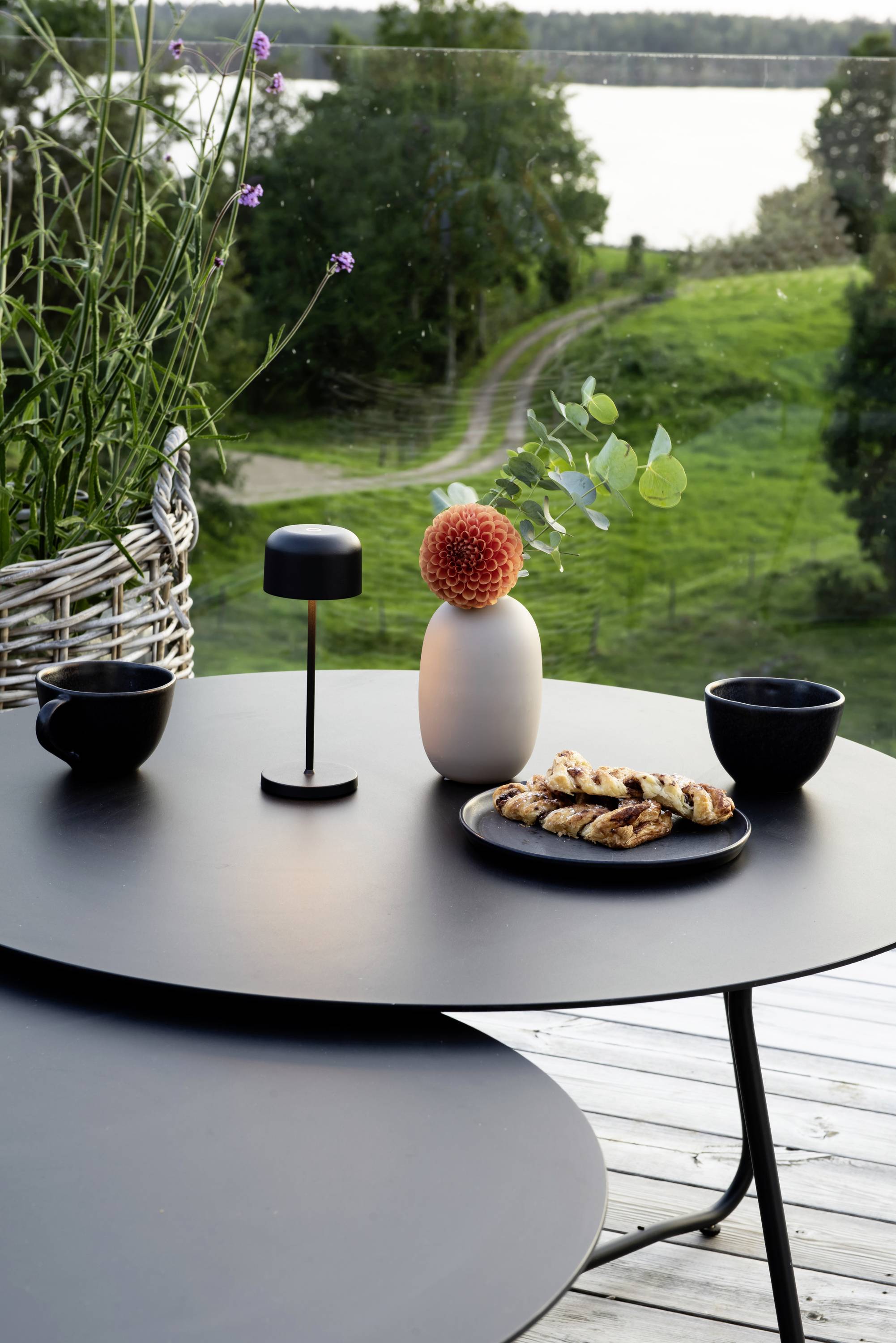 Une table ronde sur un balcon avec un couvert : un vase avec une fleur, une assiette de biscuits et deux tasses. En arrière-plan, une prairie verte et un lac.