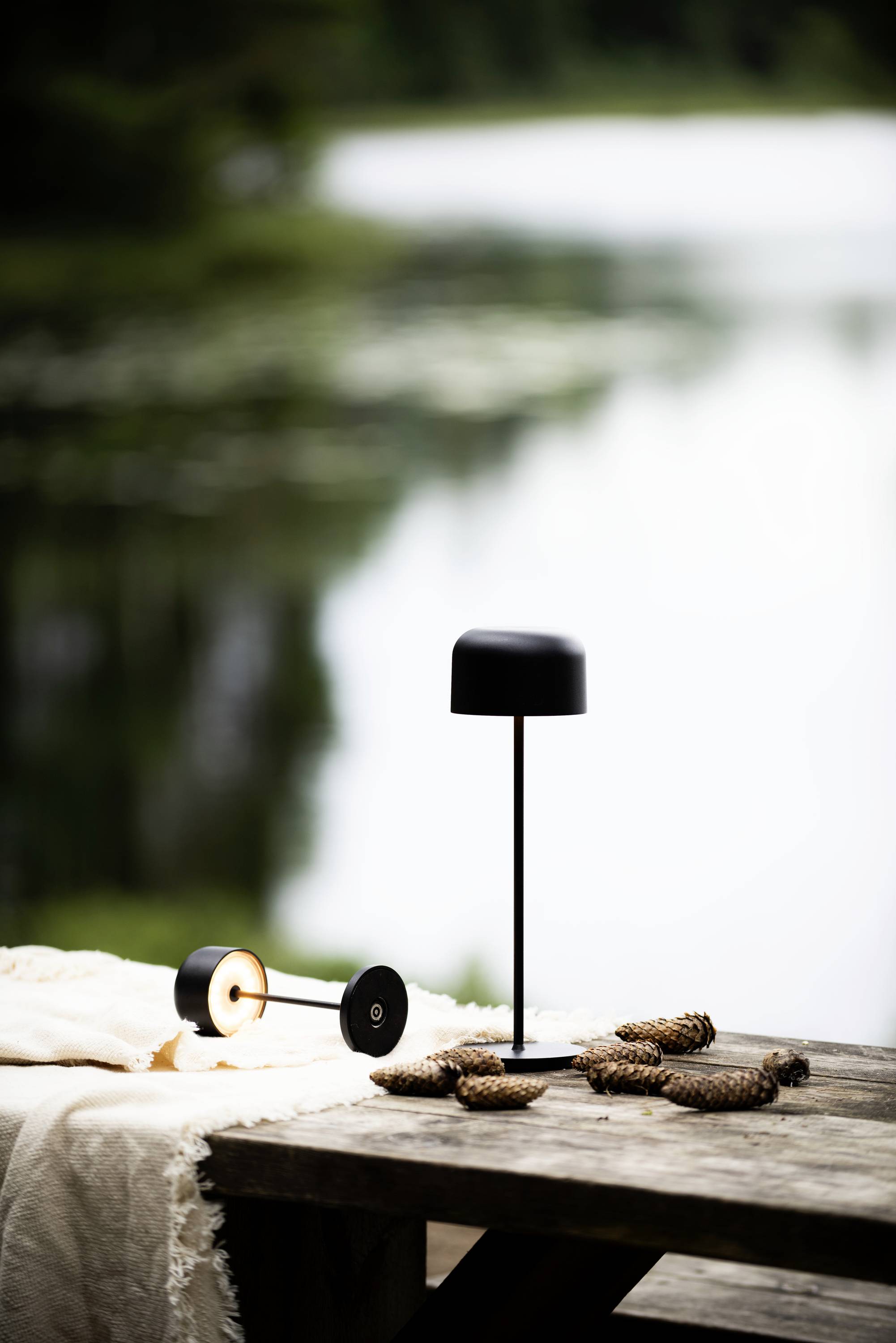 Une table à l'extérieur près du lac avec une lampe noire, des pommes de pin et un tissu beige. En arrière-plan, le lac est visible de manière floue.