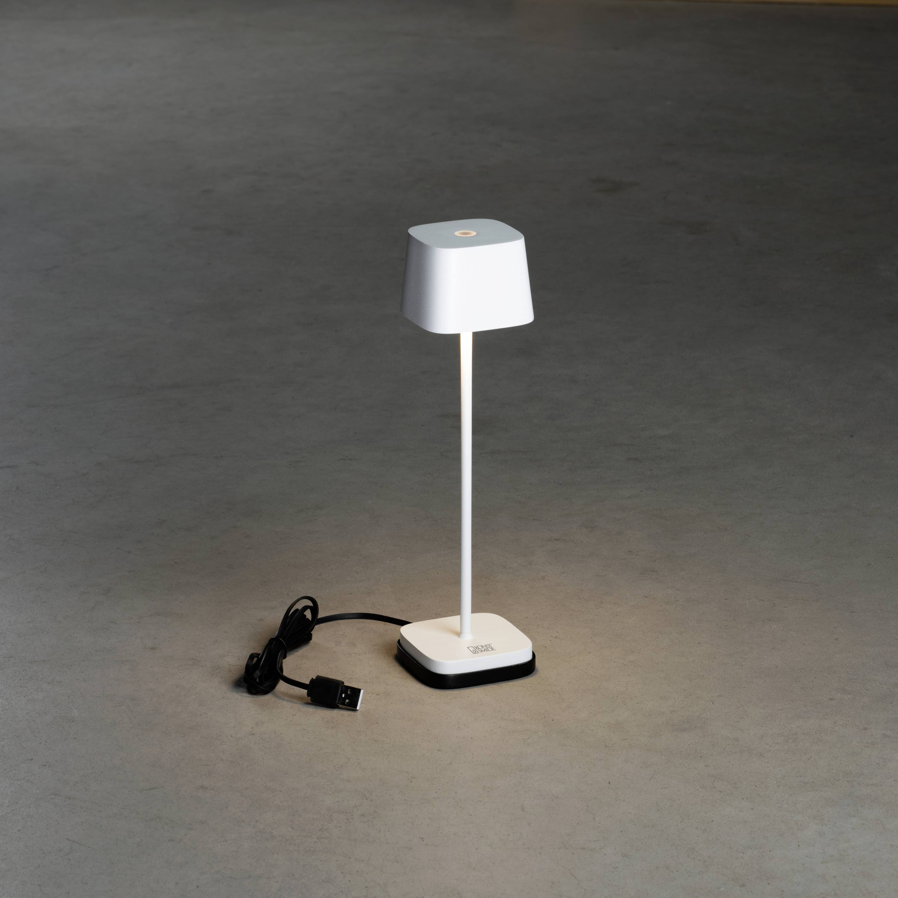 Une lampe de table simple et moderne en blanc sur un sol gris. À côté, le câble réseau noir est visible.