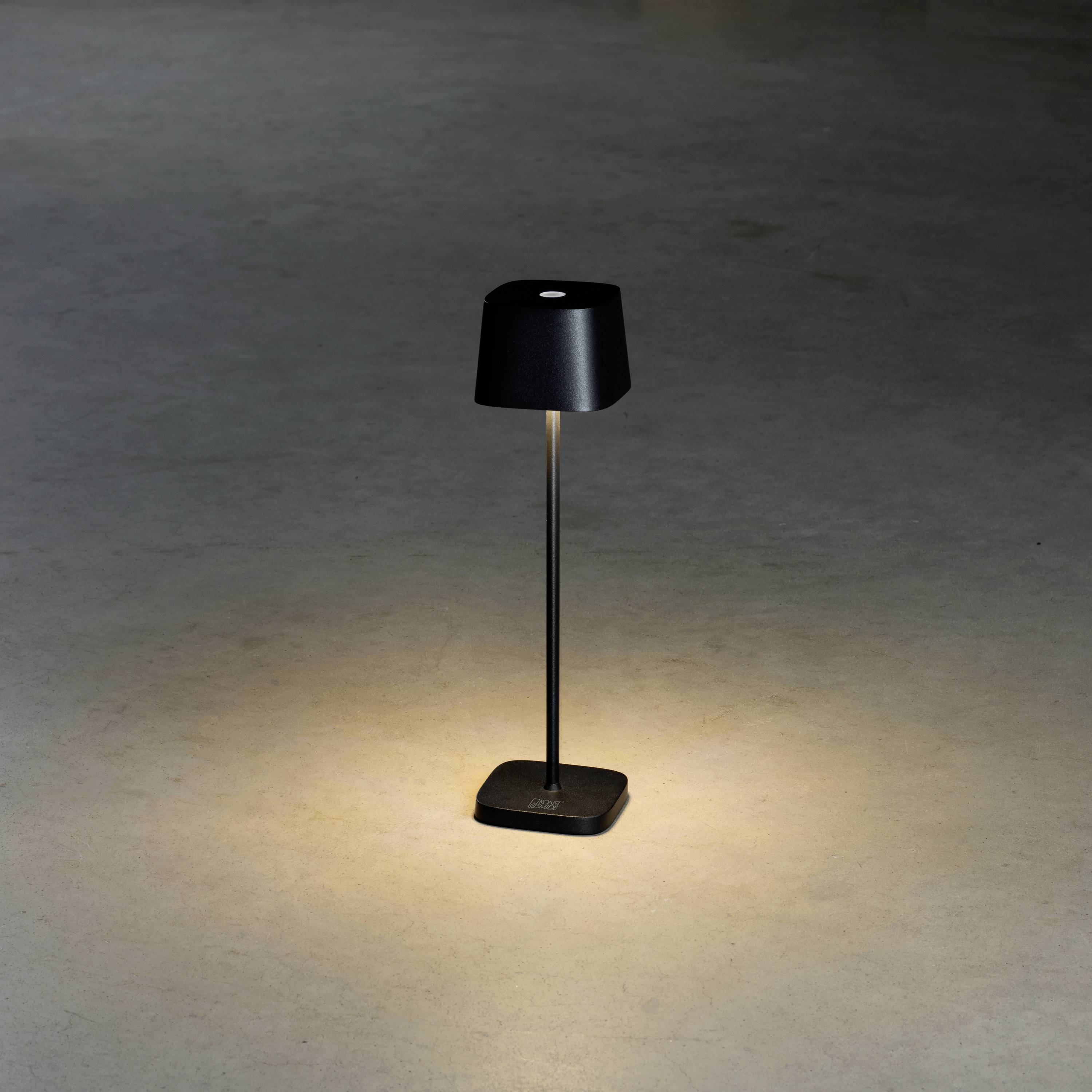 Une lampe de table noire est posée sur un sol sombre et diffuse une lumière chaleureuse sur une petite surface en dessous.