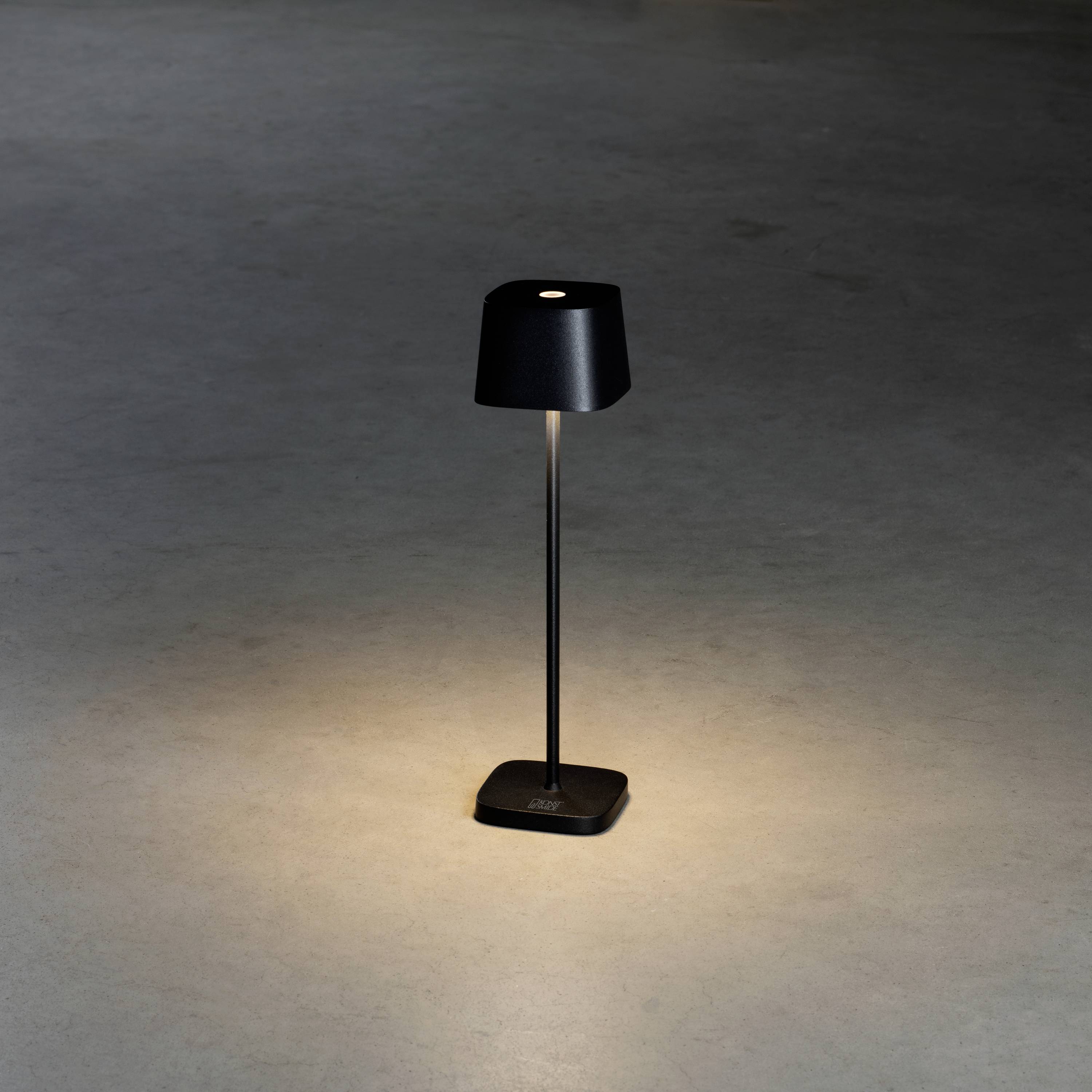 Une lampe de table noire sur un sol gris, diffusant une lumière douce vers le bas.