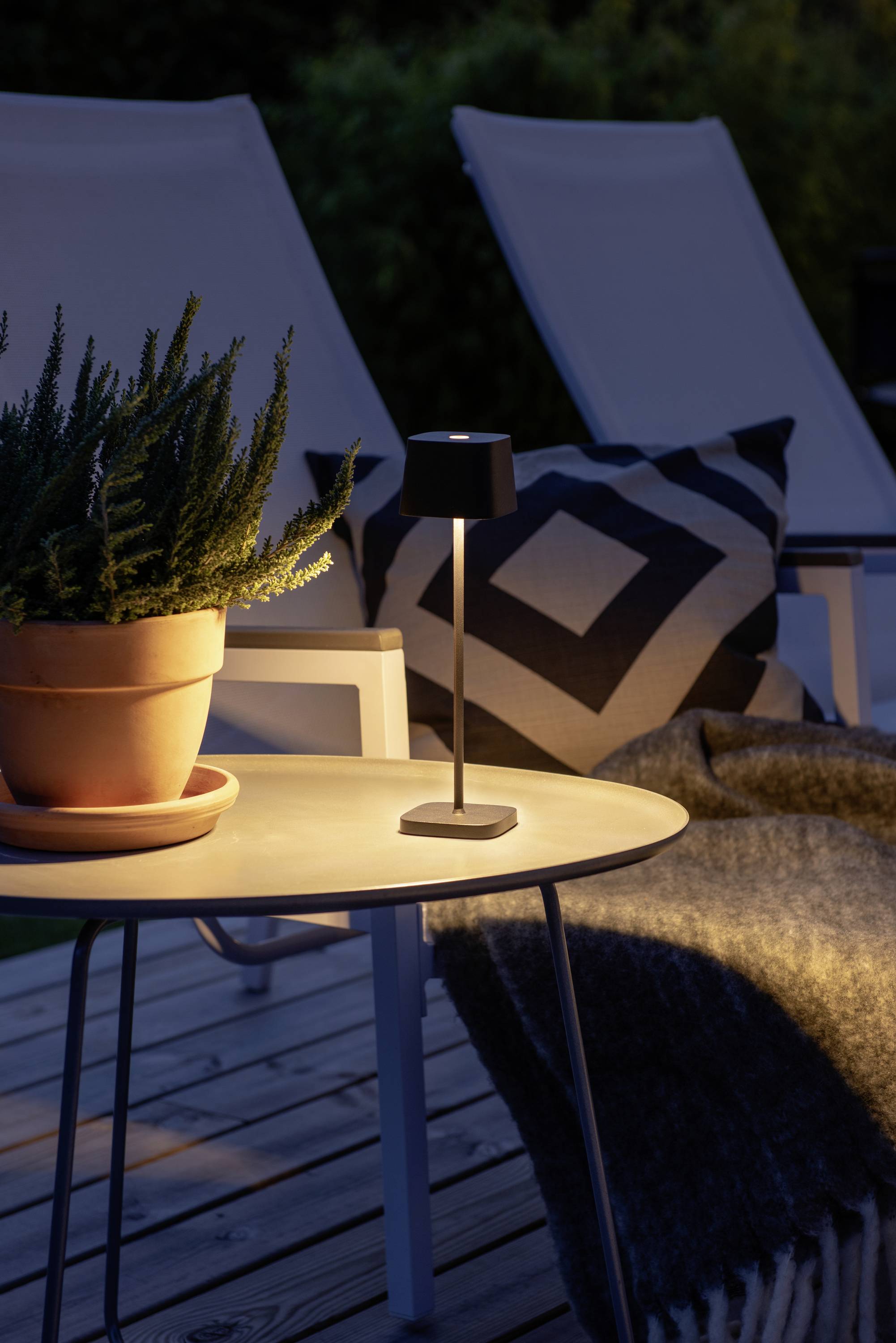 Une table éclairée doucement par une lampe illumine le salon extérieur. Dessus se trouve une plante en pot, à côté une chaise avec un coussin et une couverture.
