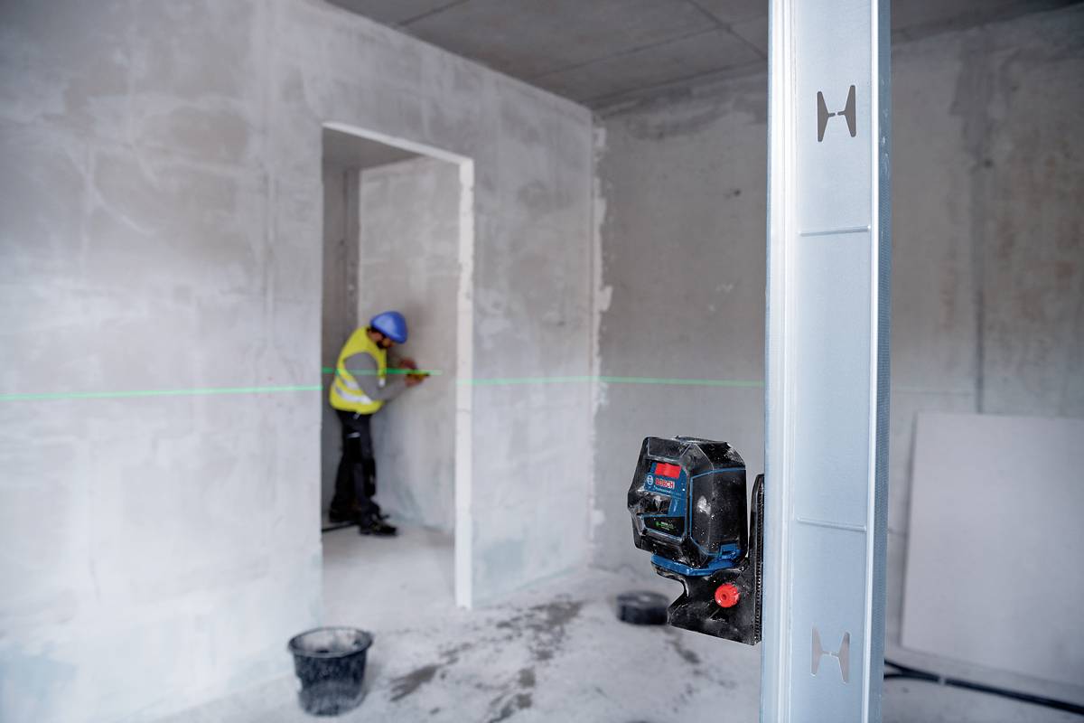 Un ouvrier portant un équipement de protection jaune mesure un mur, un appareil de nivellement laser étant installé pour effectuer les mesures.