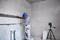 Un ouvrier portant un casque bleu aligne un mur de carrelage dans un bâtiment en construction. Un seau est posé sur le sol, à côté d'un trépied supportant un appareil laser.