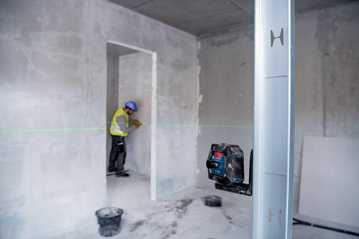 Un ouvrier en tenue de protection mesure un mur à l'aide d'un appareil laser dans une pièce inachevée. Au premier plan, un niveau laser.