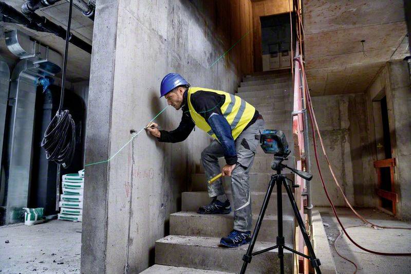 Un ouvrier portant un casque et un gilet de sécurité mesure un mur en béton sur un chantier de construction. Des équipements de construction sont visibles en arrière-plan.