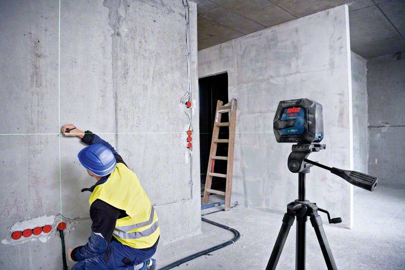 Un ouvrier marque des murs avec des traits rouges dans un bâtiment en construction. Un appareil de nivellement laser monté sur un trépied projette des lignes vertes.