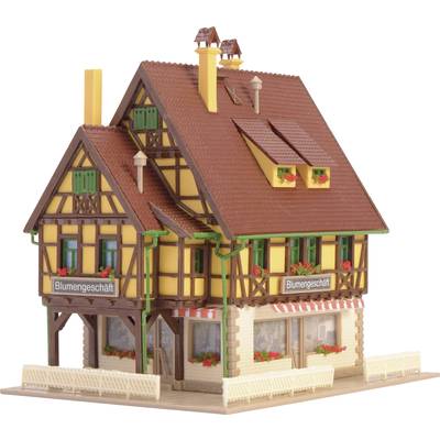 Modèle d'une maison à colombages avec l'inscription 'Magasin de fleurs'. Elle a un toit rouge, des volets verts et des jardinières.