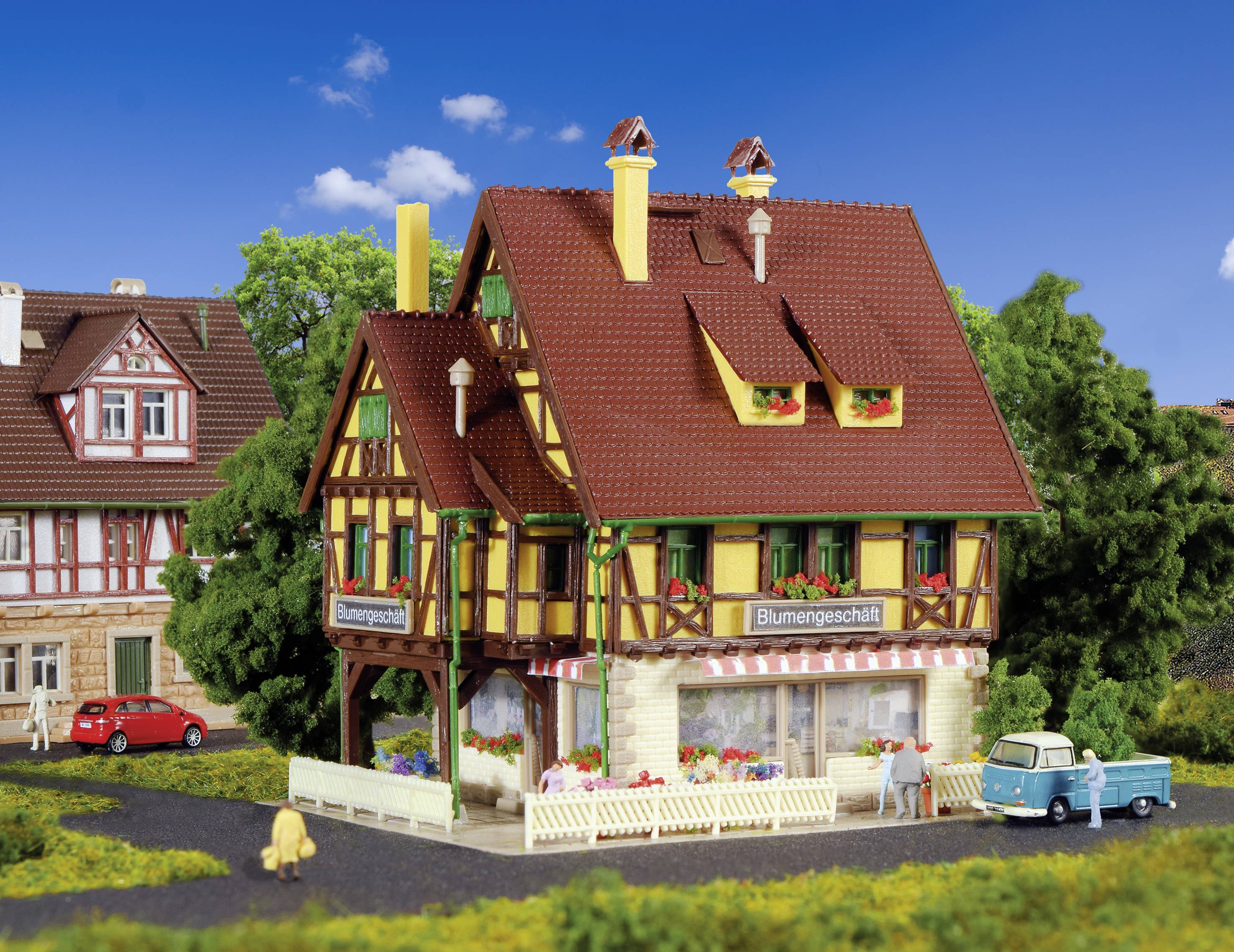 Maison à colombages avec une boutique de fleurs au rez-de-chaussée, entourée d'arbres et de voitures sur une route de campagne ; ciel ensoleillé et sans nuages.