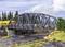 Un pont ferroviaire avec un train jaune passant à côté. En dessous, une route traverse avec deux voitures. L'arrière-plan montre des arbres et des nuages.