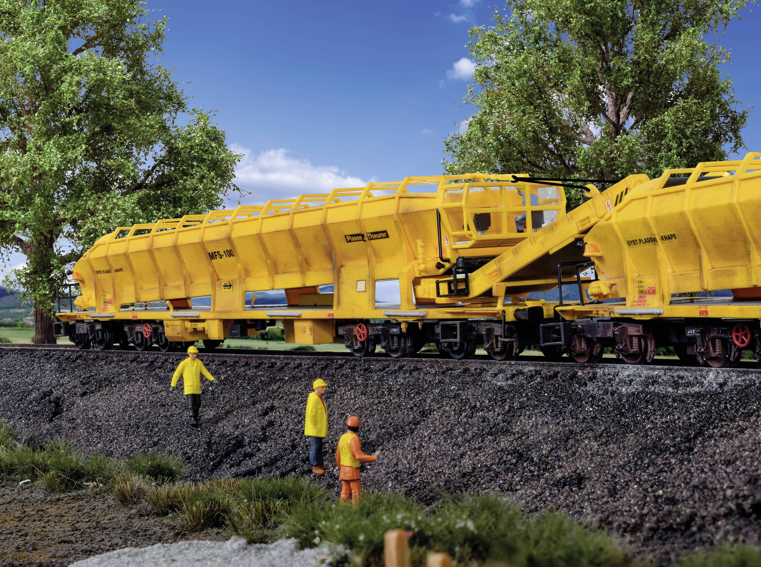 Un train de marchandises jaune avec deux ouvriers en tenue de protection sur le talus ferroviaire, entouré d'arbres et de ciel bleu.