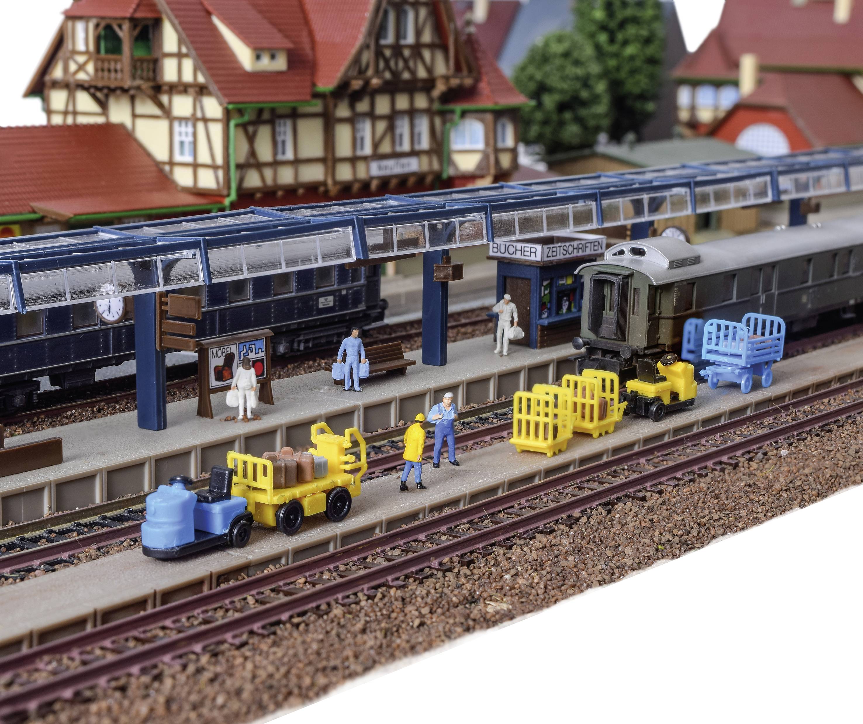 Scène de modélisme ferroviaire avec gare, train et personnages. Des ouvriers chargent des chariots de marchandises. Les bâtiments en arrière-plan créent un décor de petit village.